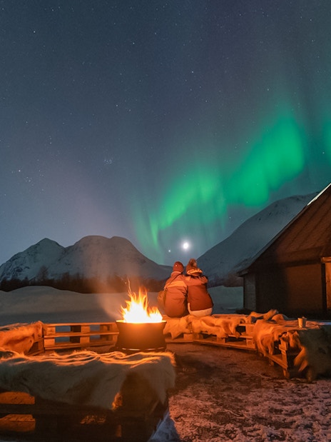 Campfire under Northern Lights with snowy mountains and cabin in the background.