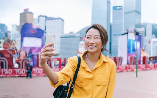 Person taking a selfie in front of Hong Kong skyline with skyscrapers.