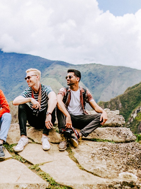 Tourists sitting on stone ledge with Machu Picchu mountains in background.