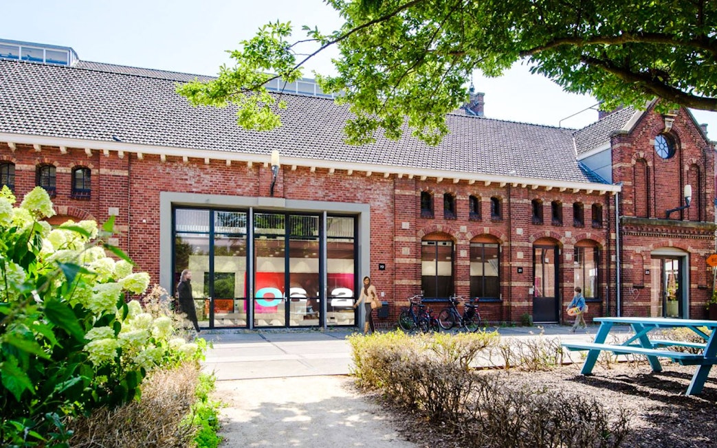 Brick building with large windows at Amsterdam in Motion exhibit, surrounded by greenery and bicycles.