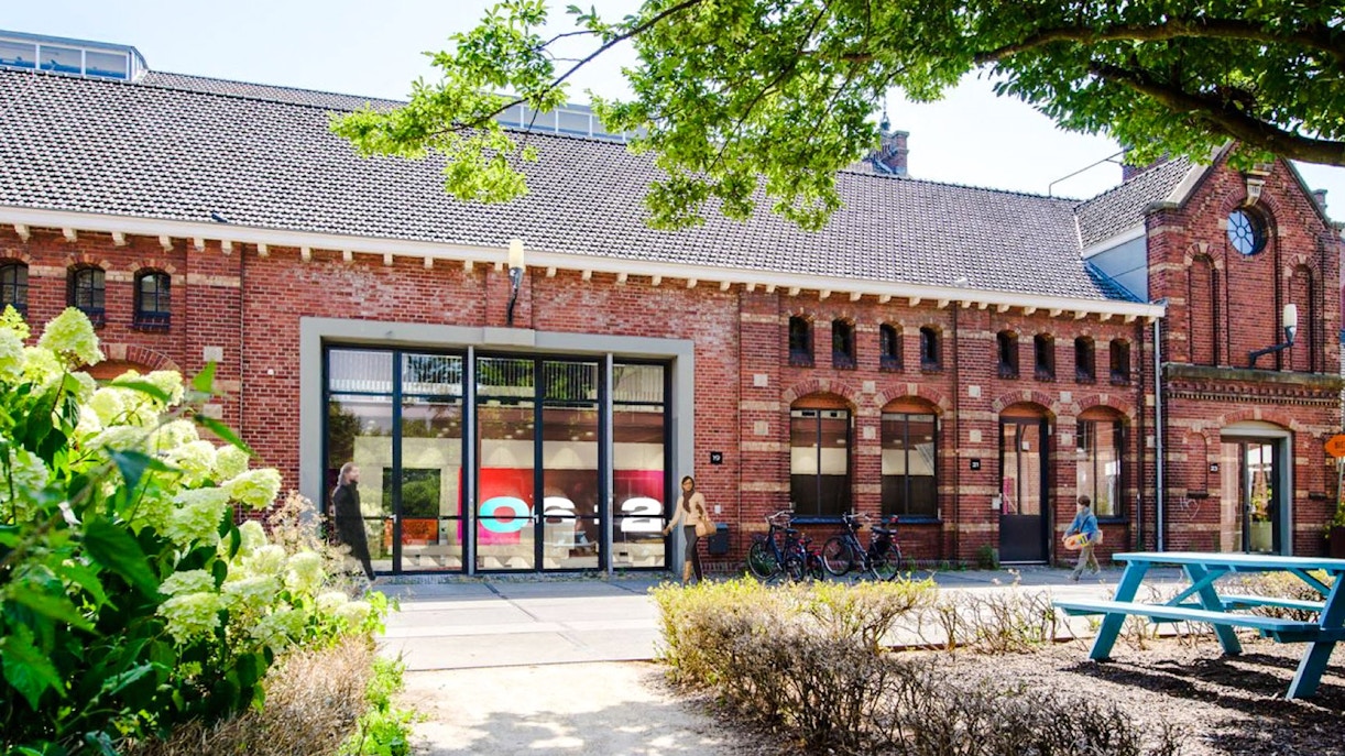 Brick building with large windows at Amsterdam in Motion exhibit, surrounded by greenery and bicycles.