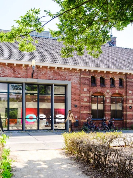 Brick building with large windows at Amsterdam in Motion exhibit, surrounded by greenery and bicycles.