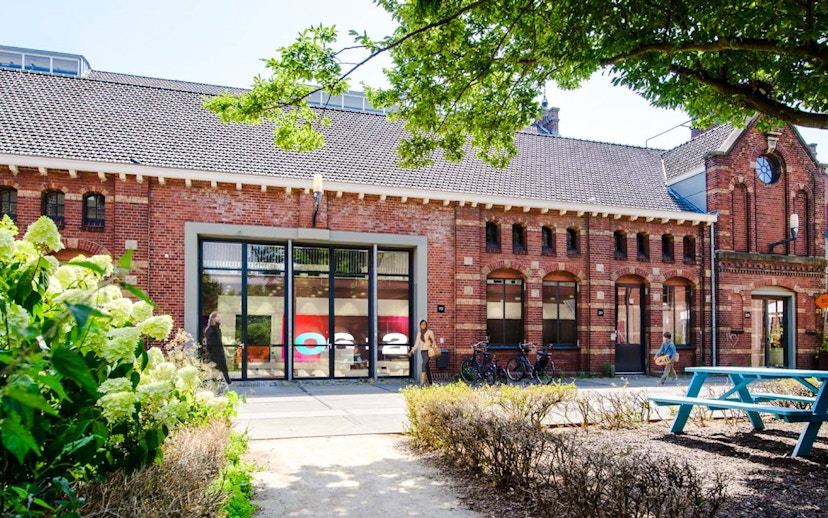 Brick building with large windows at Amsterdam in Motion exhibit, surrounded by greenery and bicycles.