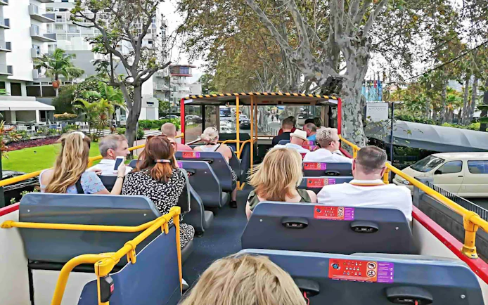People seated on upper deck of City Sightseeing bus in Funchal, passing by trees and buildings.