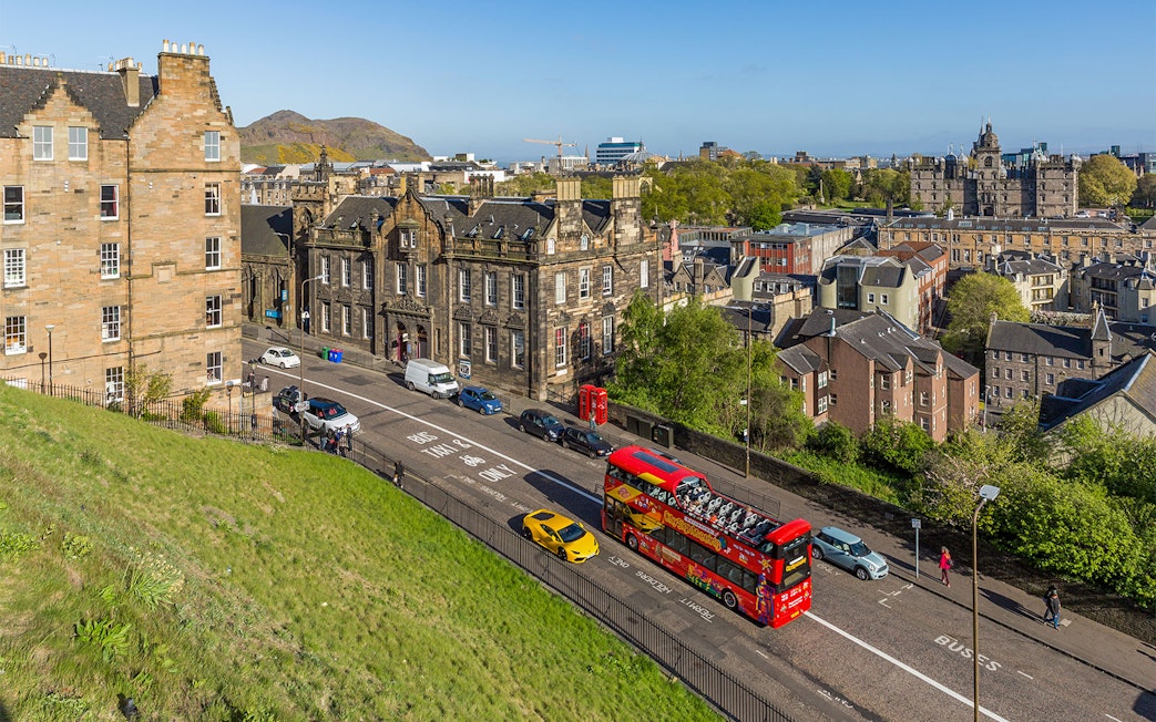 Hop-On Hop-Off bus on a street in Edinburgh with historic buildings and Arthur's Seat in the background.