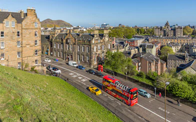 Hop-On Hop-Off bus on a street in Edinburgh with historic buildings and Arthur's Seat in the background.