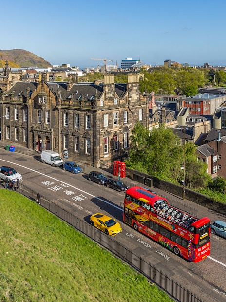 Hop-On Hop-Off bus on a street in Edinburgh with historic buildings and Arthur's Seat in the background.