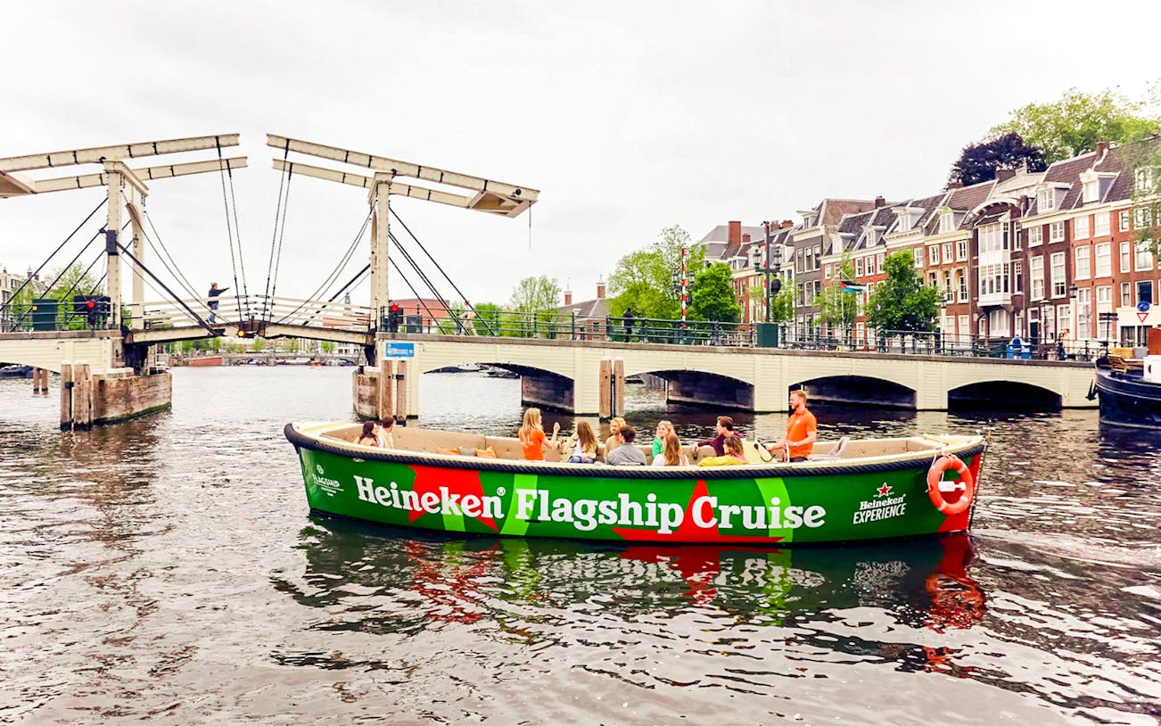 Tourists enjoying Heineken Experience flagship cruise on Amsterdam canal with cityscape views.