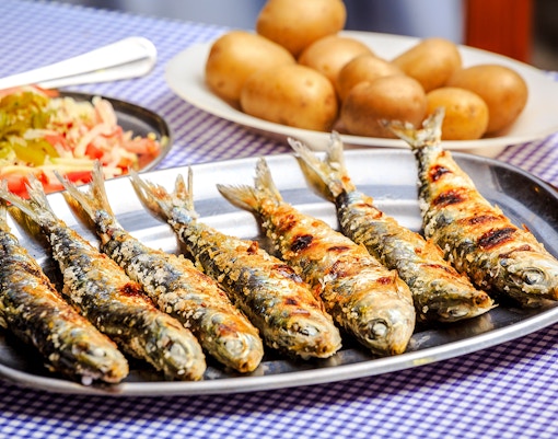 Grilled sardines on a plate with potatoes and salad in the background.