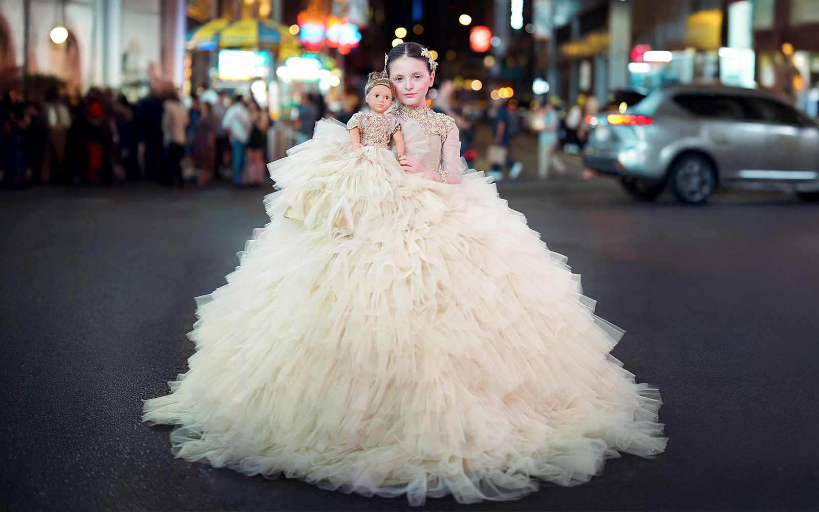 Girl in a formal dress holding a matching doll on a city street at night.