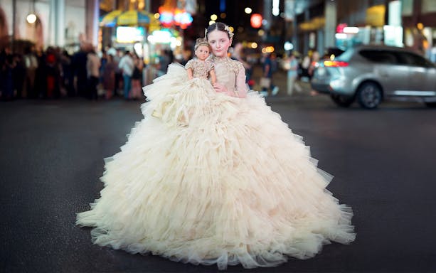 Girl in a formal dress holding a matching doll on a city street at night.