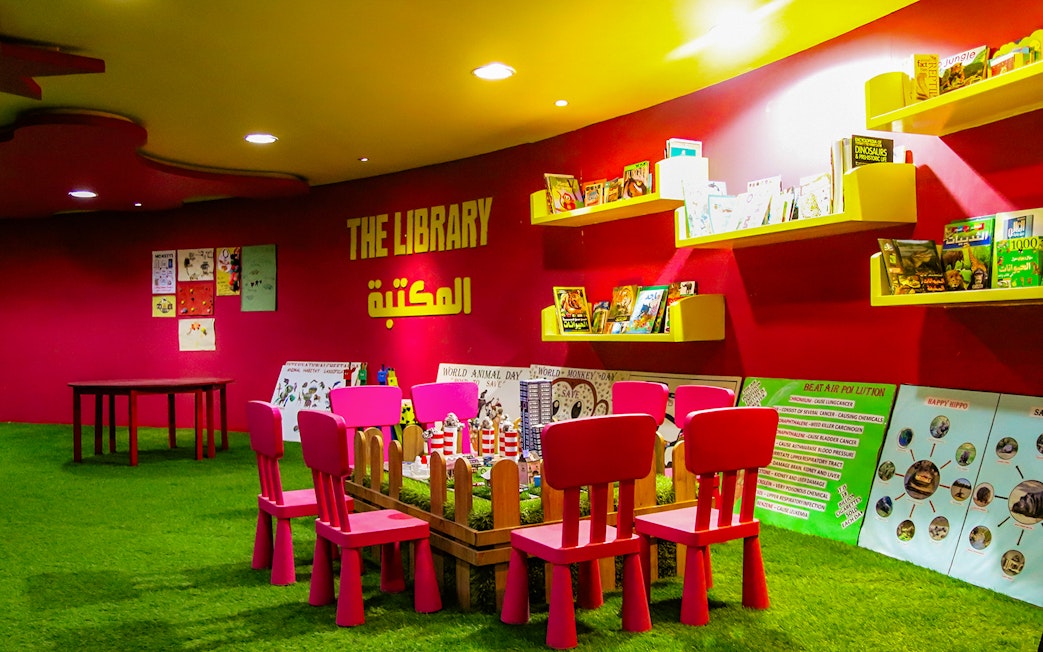 Children's reading area in a colorful library with books on shelves and small chairs around a table.