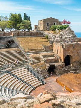 Ancient Roman theater in Catania with stone seating and stage, surrounded by ruins and trees.
