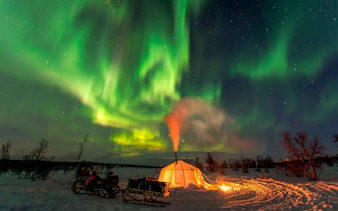Northern lights over a lit tent in snowy Abisko landscape.
