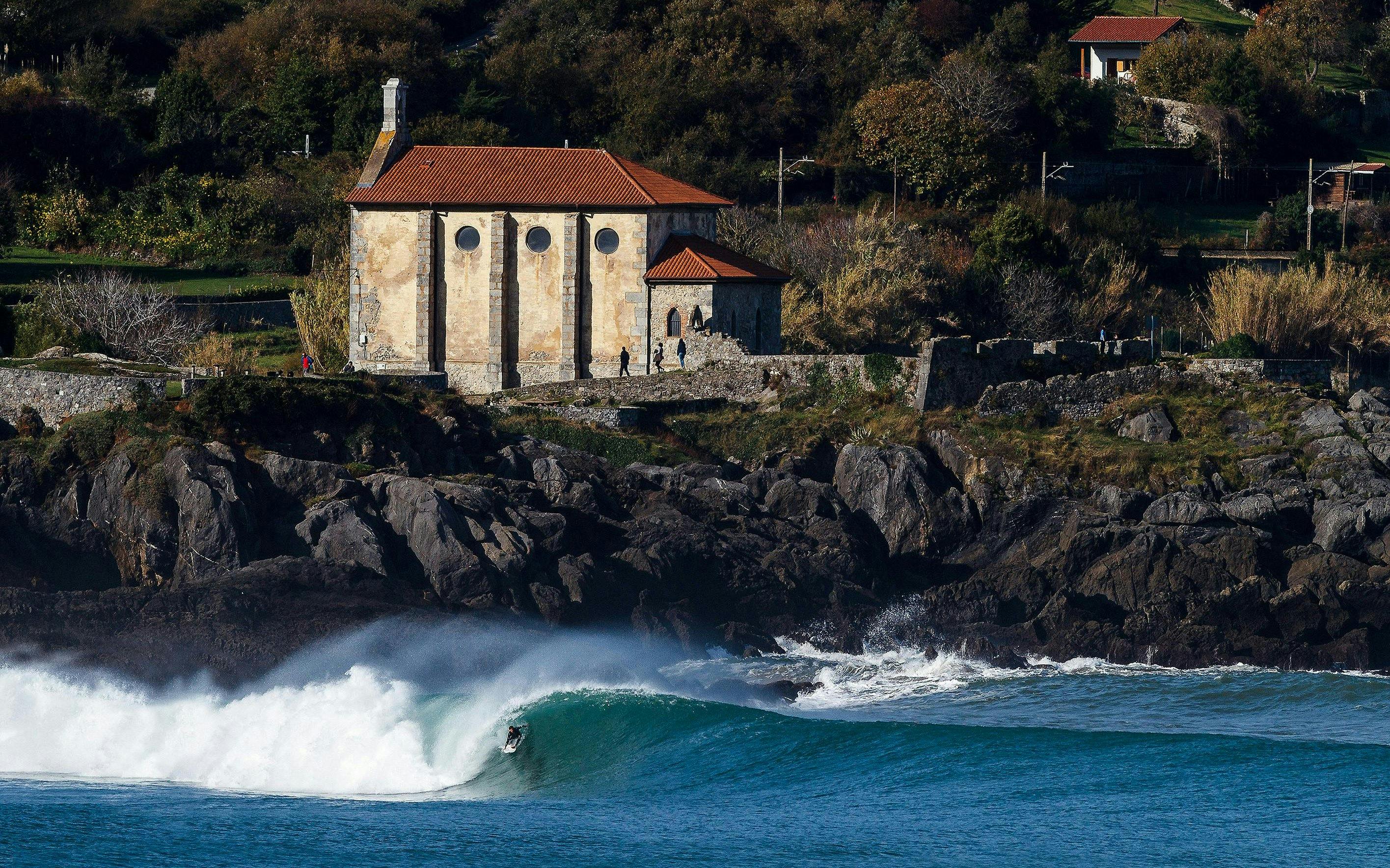 Surfer riding a wave near a historic building in Mundaka, Spain.