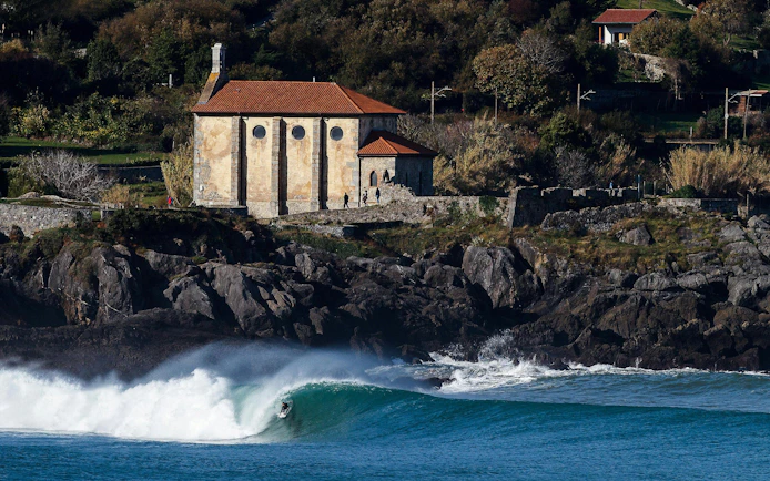 Surfer riding a wave near a historic building in Mundaka, Spain.