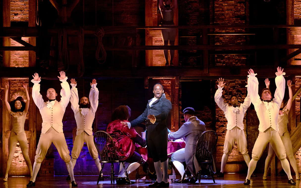 Stage performance of Hamilton with actors in period costumes, featuring a central figure addressing the audience.