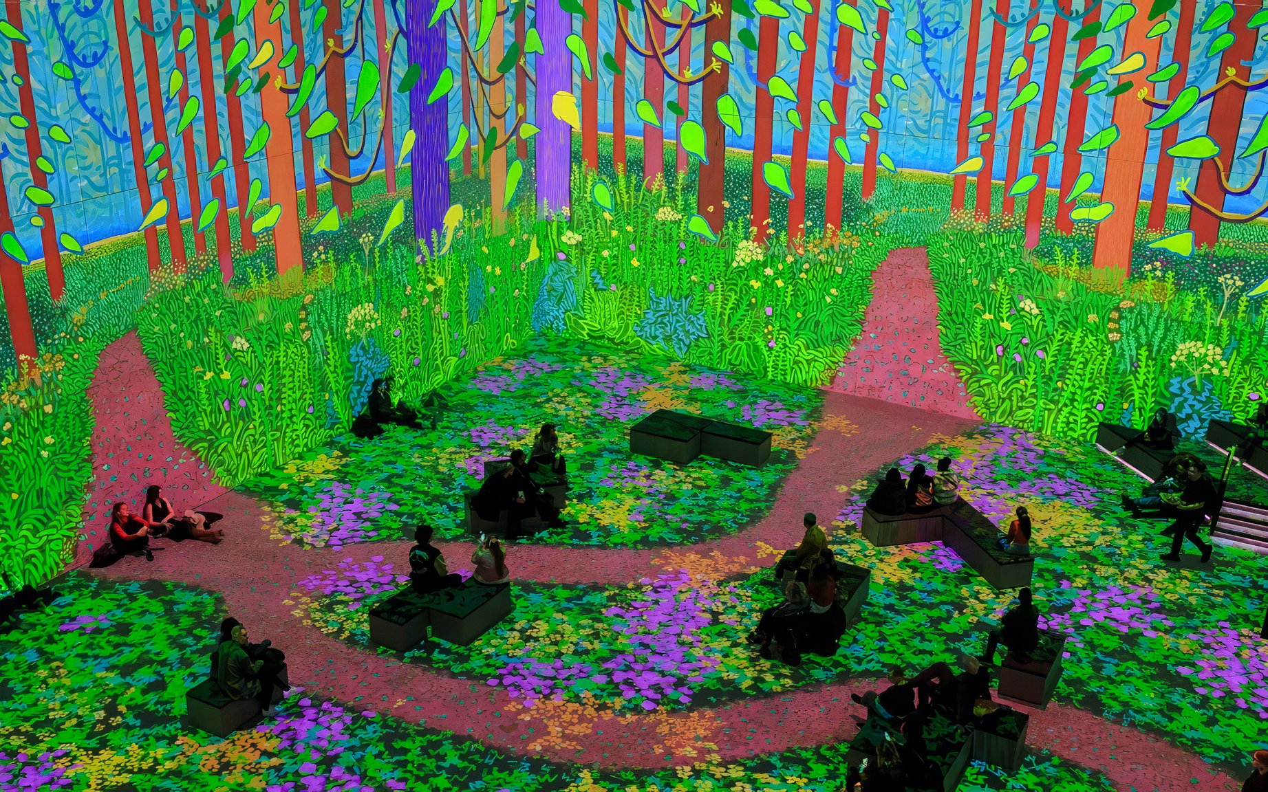 Immersive digital forest scene from David Hockney: Bigger & Closer exhibition with seated visitors.