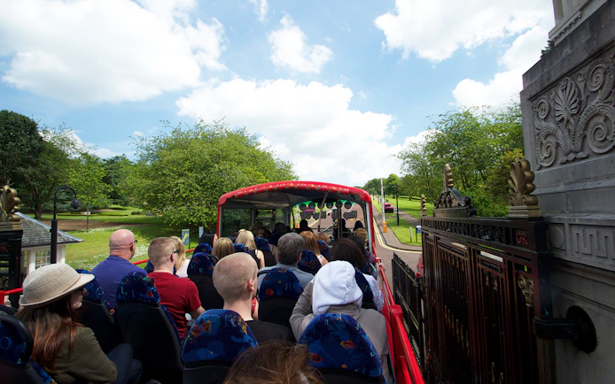 Open-top bus with tourists on Belfast Hop-On Hop-Off Tour passing through a park.