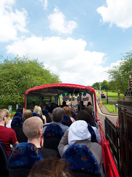 Open-top bus with tourists on Belfast Hop-On Hop-Off Tour passing through a park.