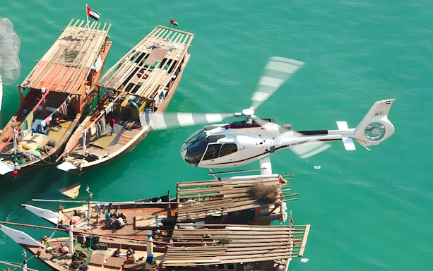 Helicopter flying over traditional boats on Dubai's turquoise waters.