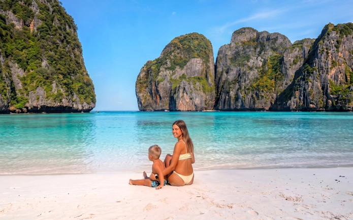 Woman and child sitting on Phi Phi Island beach with limestone cliffs and turquoise water in the background.
