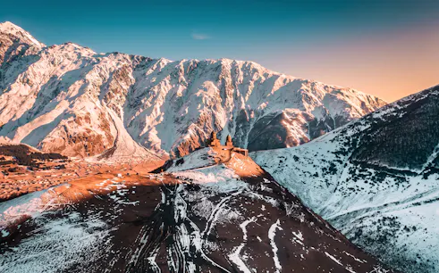 Gergeti Trinity Church on a snowy mountain in Georgia with a sunset backdrop.