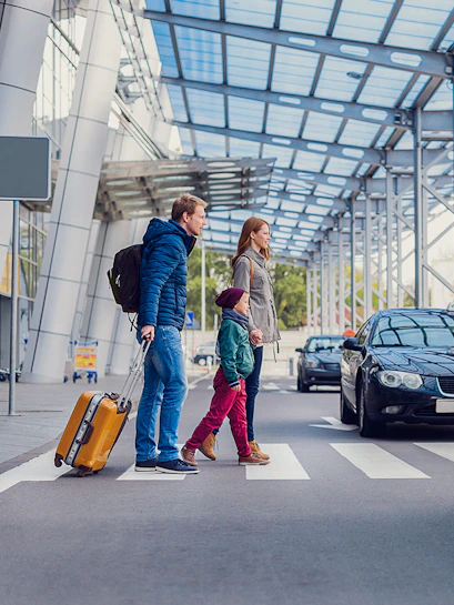 Family crossing road at airport with parked taxis nearby.