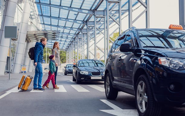 Family crossing road at airport with parked taxis nearby.