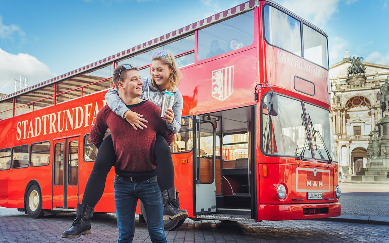 Couple enjoying Dresden bus tour with live commentary in front of red double-decker bus.
