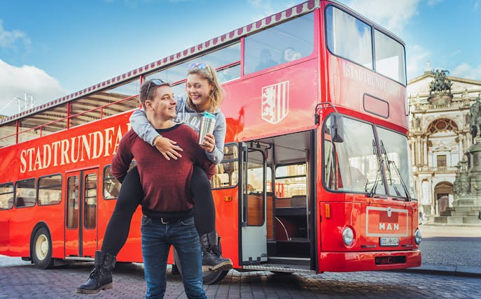 Couple enjoying Dresden bus tour with live commentary in front of red double-decker bus.