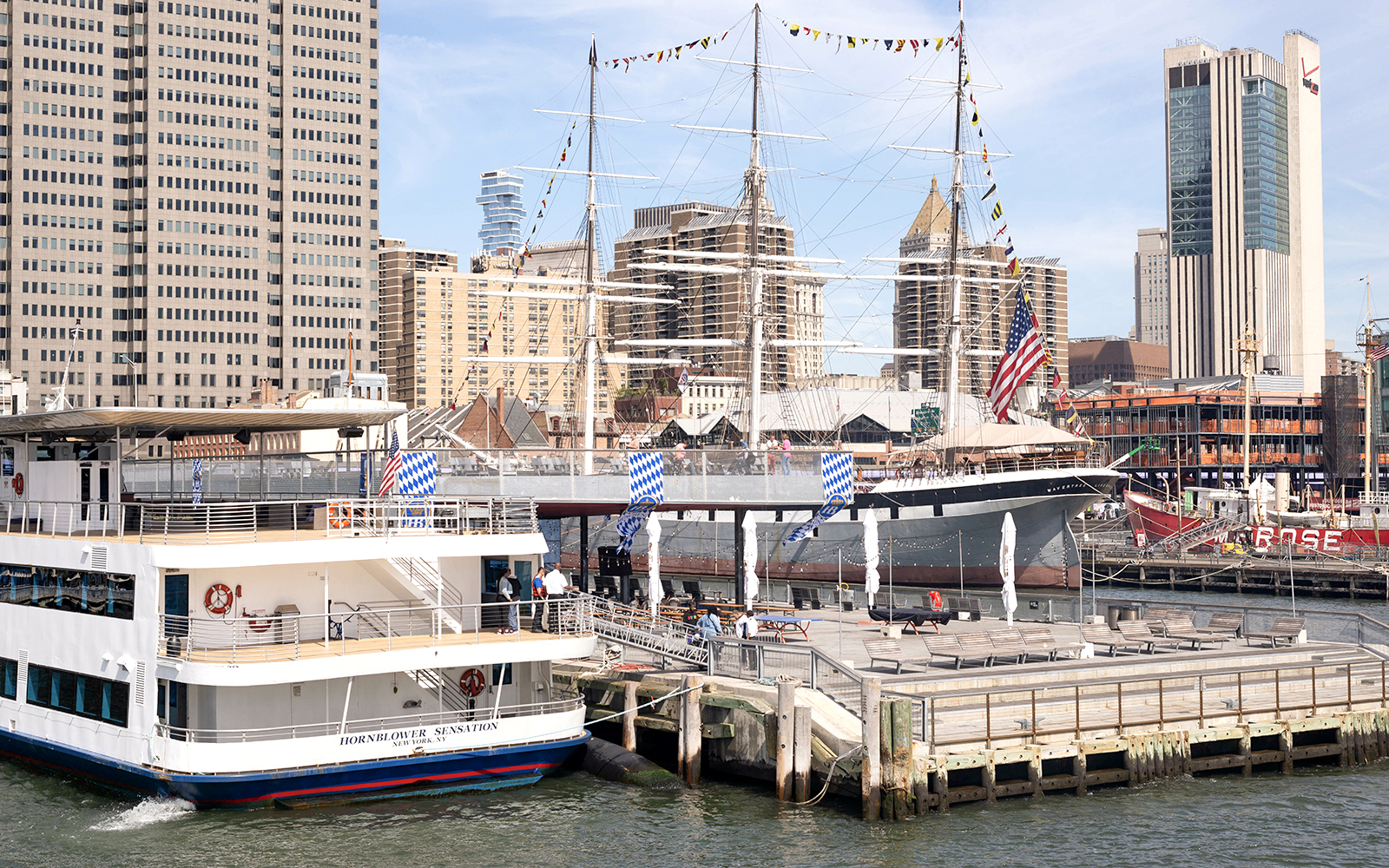 Tour boat docked at South Street Seaport with historic ship and New York City skyline.