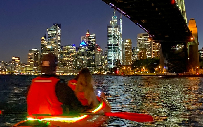 Couple kayaking under Sydney Harbour Bridge at night with city skyline.