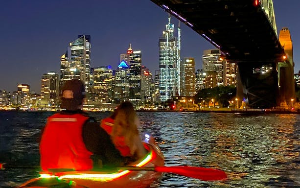 Couple kayaking in LED-lit kayak under Sydney Harbour Bridge at night with city lights in the background.