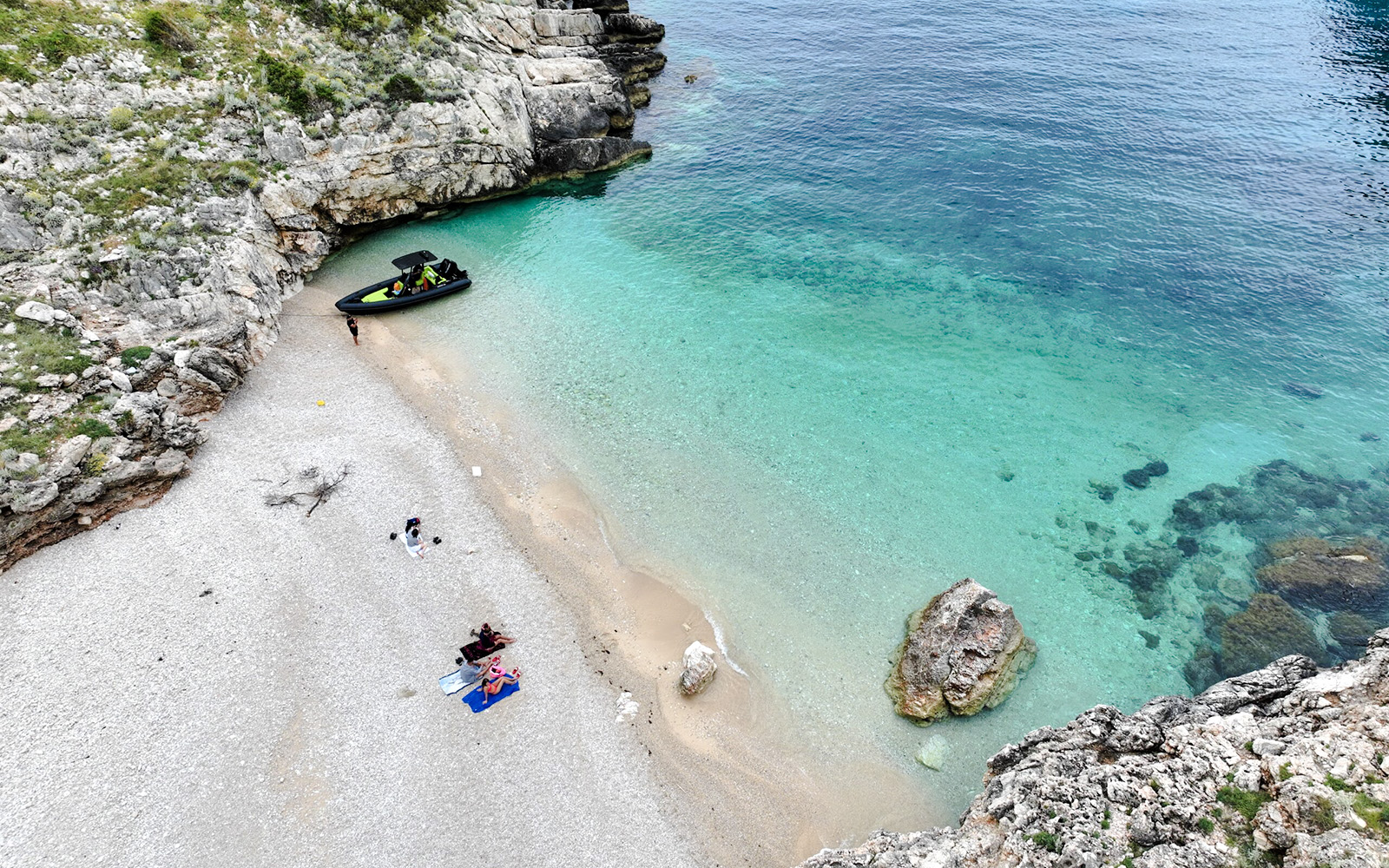 Small group relaxing on Dafina Bay beach near Haxhi Ali Cave with boat in turquoise water.