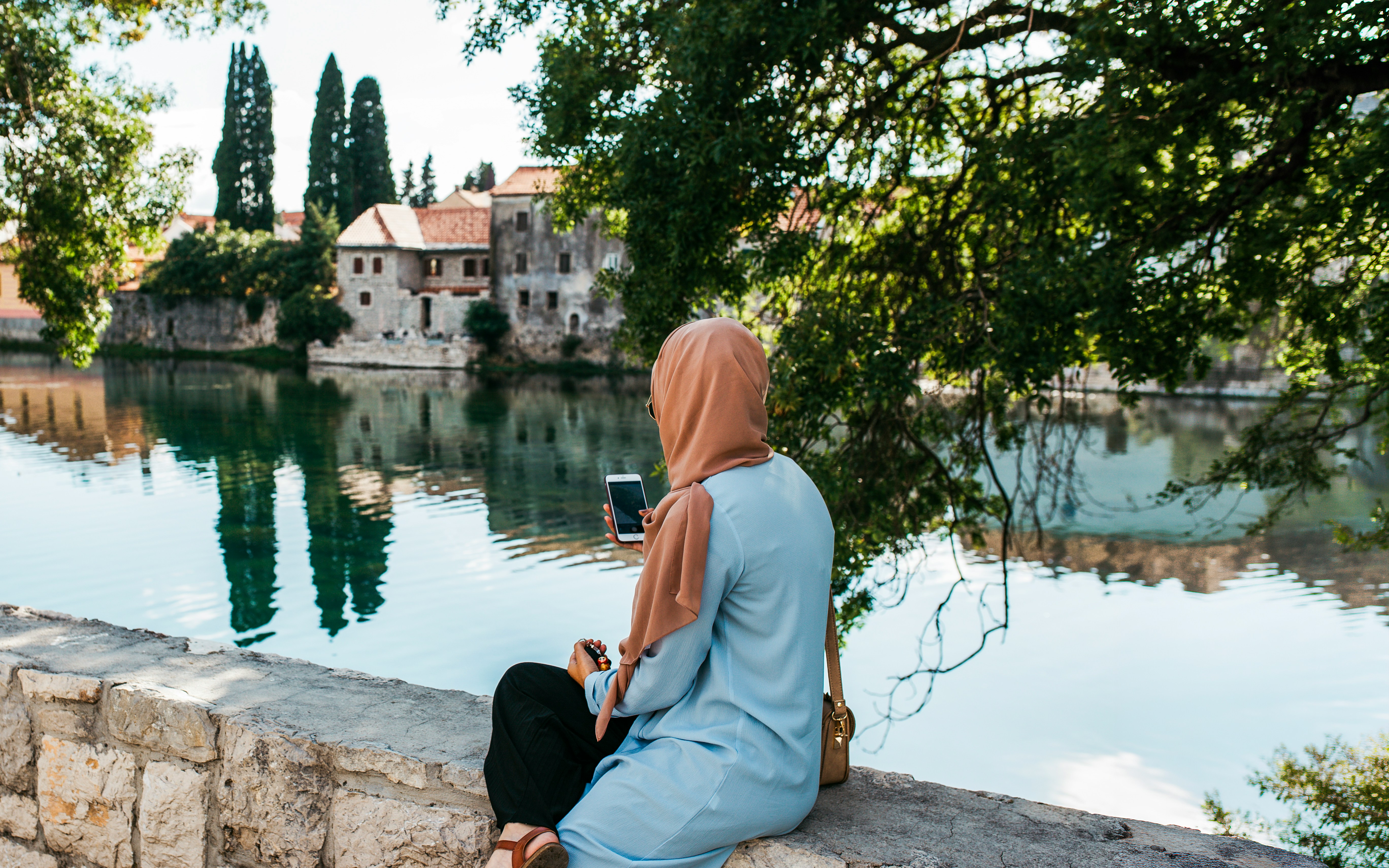 Woman sitting by Trebisnjica River in Trebinje, Bosnia and Herzegovina, with historic buildings.