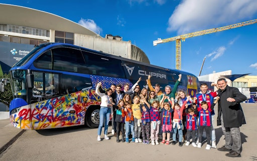 Group of people posing in front of a colorful Barça team bus at the J. Cruyff Stadium.