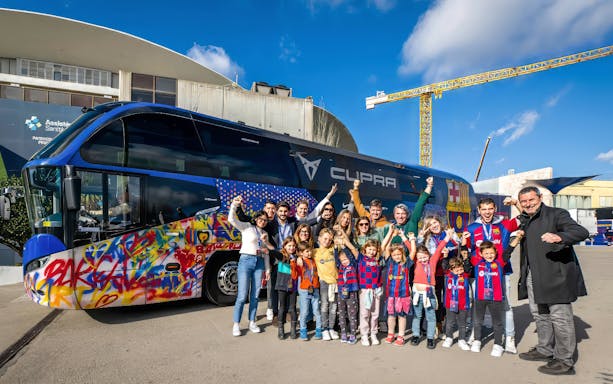 Group of people posing in front of a colorful Barça team bus at the J. Cruyff Stadium.