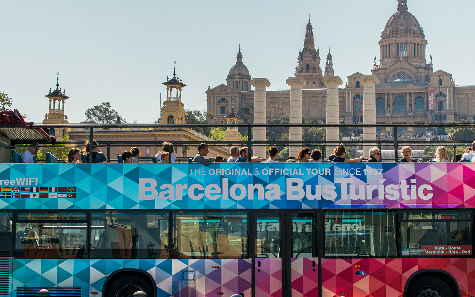 Barcelona Bus Turistic with tourists, Montjuïc Palace in background.