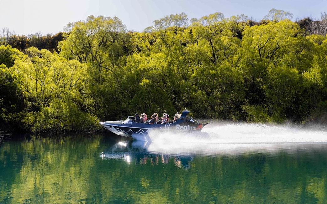 Jet boat speeding on Kawarau River with passengers, surrounded by lush greenery.