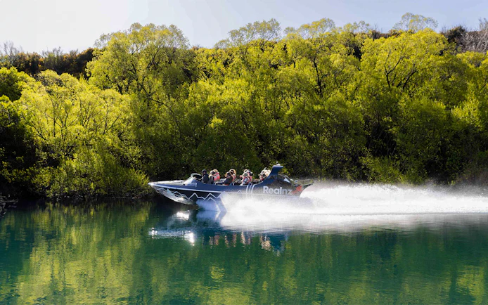 Jet boat speeding on Kawarau River with passengers, surrounded by lush greenery.