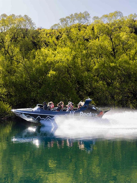 Jet boat speeding on Kawarau River with passengers, surrounded by lush greenery.