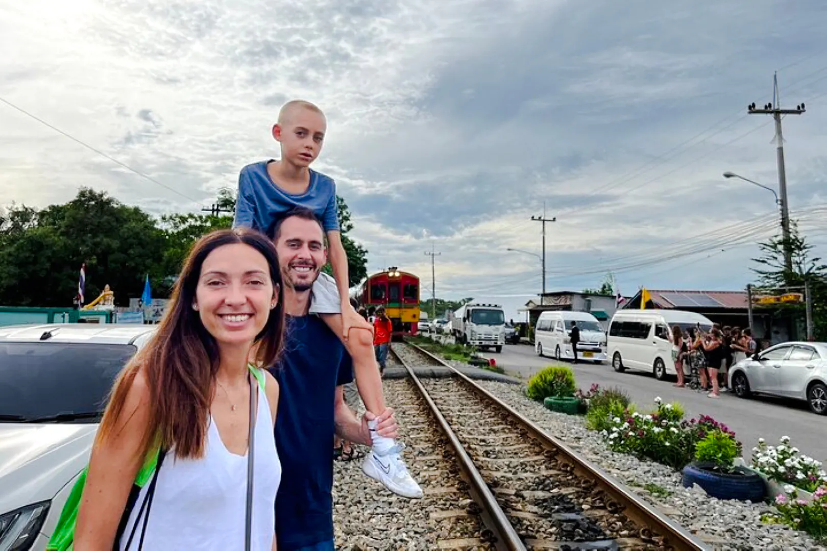 Family standing by railway tracks with train approaching at Railway Market, Thailand.