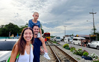 Family standing by railway tracks with train approaching at Railway Market, Thailand.