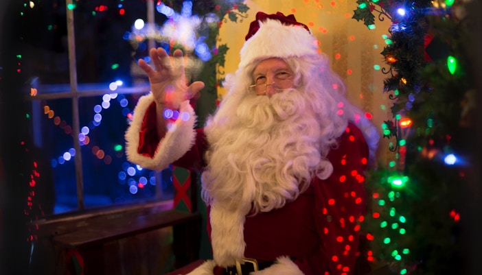 Santa Claus waving in a festive room with colorful lights.