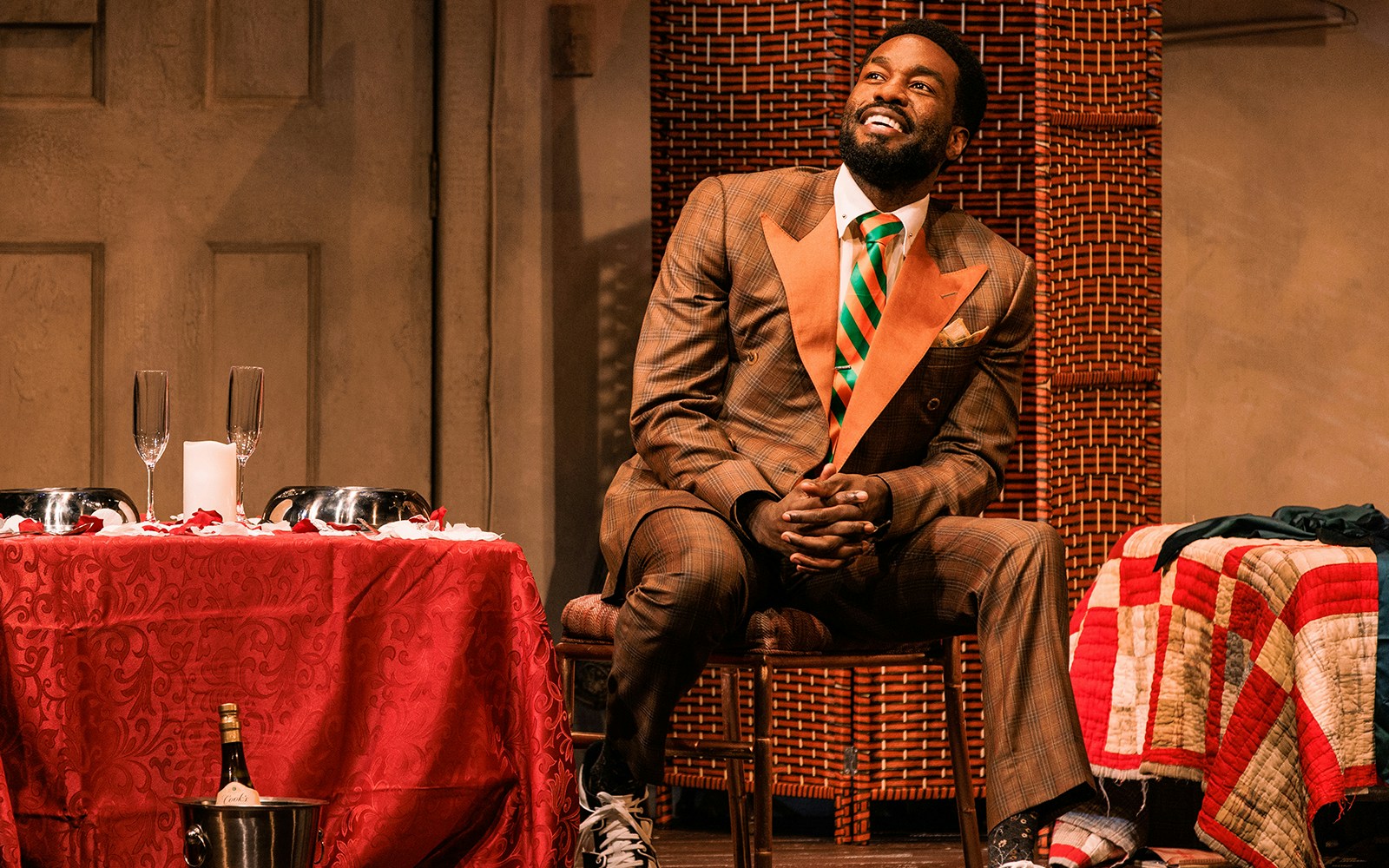 Man in a suit sitting beside a table with champagne glasses and a red tablecloth.