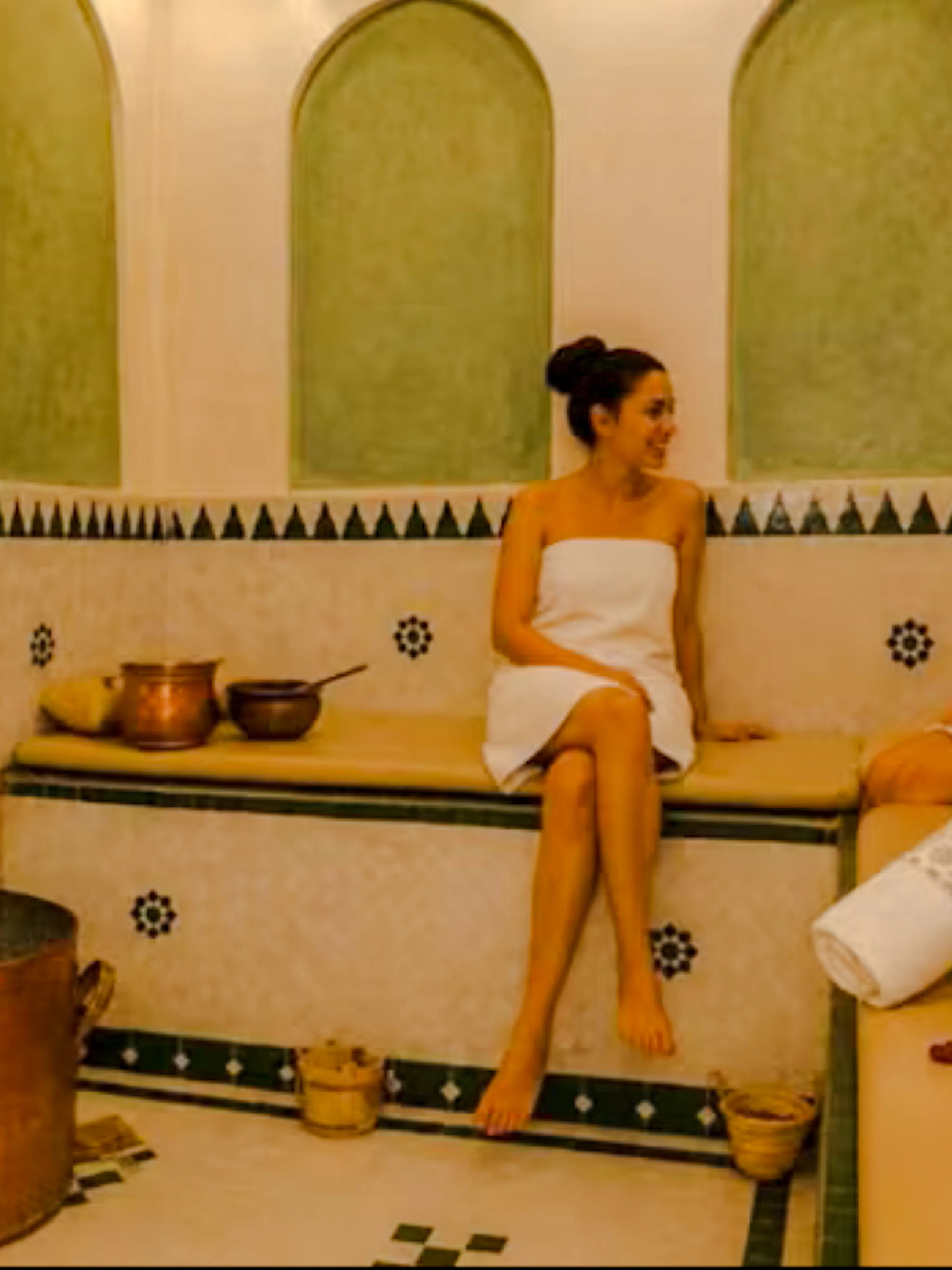 Two women in a traditional Moroccan hammam in Marrakech, surrounded by copper pots and towels.
