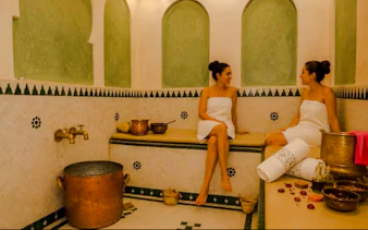 Two women in a traditional Moroccan hammam in Marrakech, surrounded by copper pots and towels.