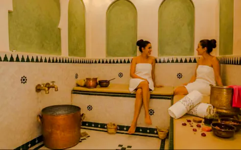 Two women in a traditional Moroccan hammam in Marrakech, surrounded by copper pots and towels.