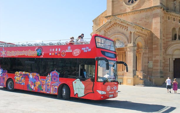 Red double-decker bus for Gozo Hop On Hop Off tour near a historic church.